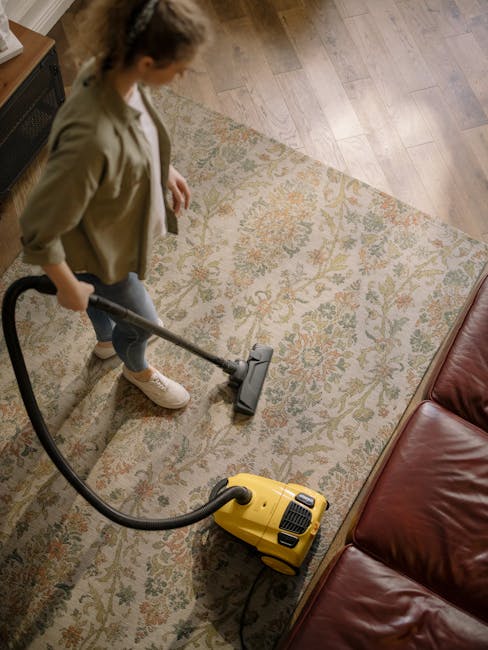 A close-up view of a person vacuuming a traditional patterned area rug with a black and grey upright vacuum cleaner, which is positioned on a well-maintained wooden floor adjacent to the rug. The pers